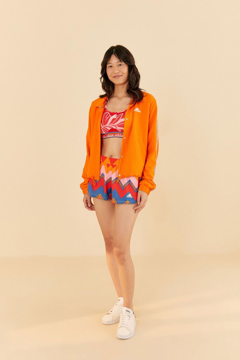 Modelo vistiendo el short Adidas estampado con patrón geométrico vibrante, combinado con chaqueta naranja y zapatillas blancas.