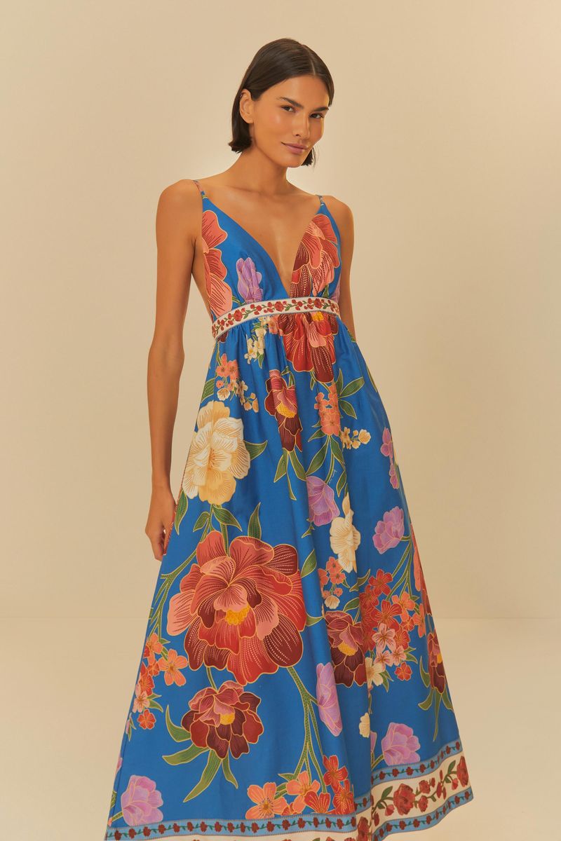 Modelo usando un vestido largo estampado con tirantes delgados y escote profundo, fondo azul vibrante con flores coloridas.