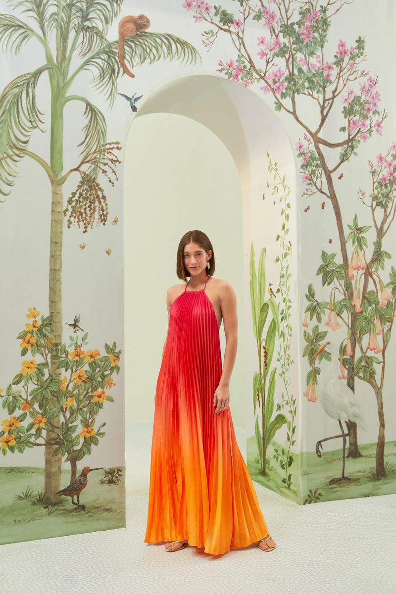 Vestido largo plisado con efecto degradado en tonos rojo y naranja, visto desde el frente en ambiente decorado.