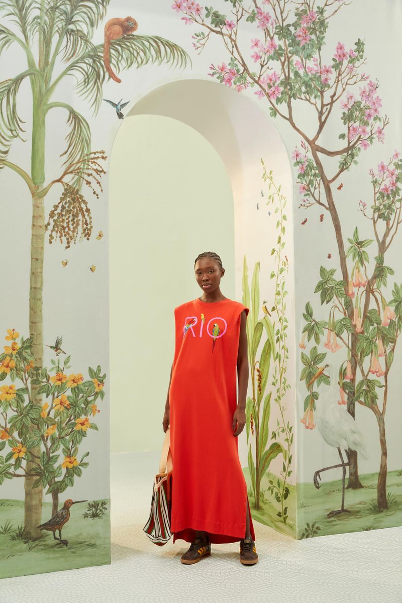 Vestido largo rojo con bordado 'RIO' mostrado en un entorno decorado con elementos tropicales.