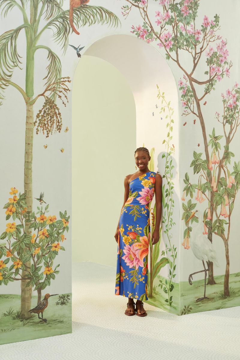 Modelo usando vestido midi tropical con estampado de flores y frutas en fondo azul, posando en escenario con plantas.
