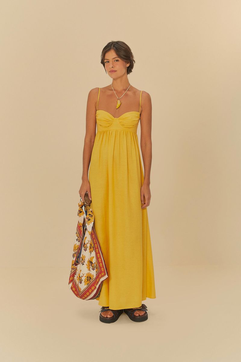 Imagen de un vestido largo amarillo con tirantes finos, modelo sosteniendo un bolso estampado.