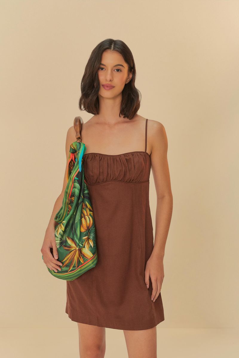 Imagen de una mujer usando un vestido corto fruncido marrón con tirantes finos, sosteniendo una bolsa estampada verde.
