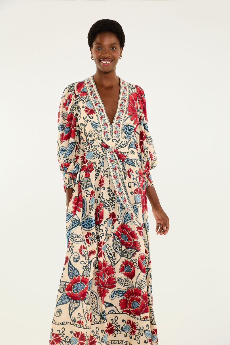 Imagen de un vestido largo con mangas abullonadas y estampado floral en tonos de rojo y azul sobre fondo beige, visto de lado.