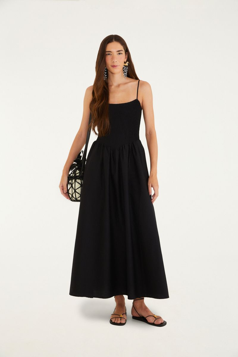 Imagen de una mujer vistiendo un vestido negro largo con tirantes delgados, sosteniendo una bolsa negra y usando sandalias.