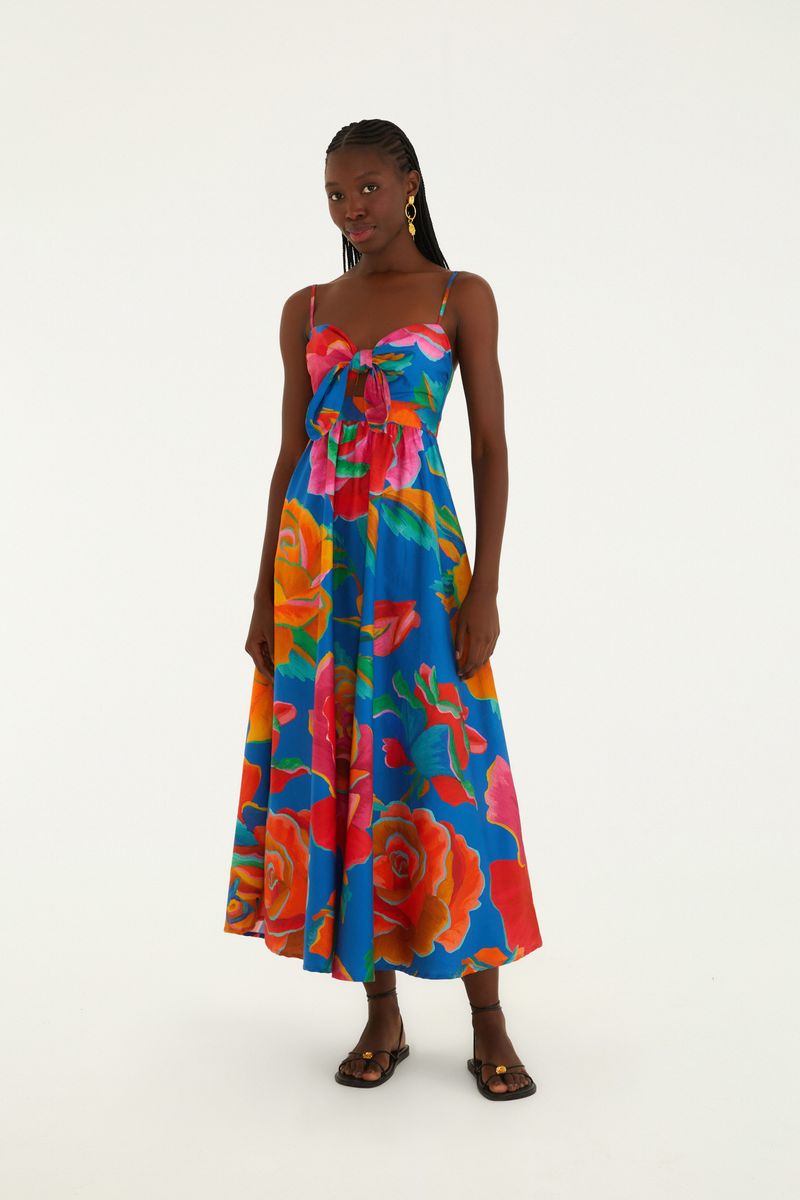 Vestido Marias Rosas Pintadas: modelo de pie usando vestido largo azul con estampado floral sobredimensionado de rosas en tonos de rosa, naranja y verde, tirantes finos, nudo frontal y sandalias planas.