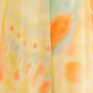 Primer plano del tejido del pantalón Asas: estampado tie-dye de acuarela con grandes manchas naranjas, puntos azules sobre fondo amarillo claro, textura ligera y translúcida